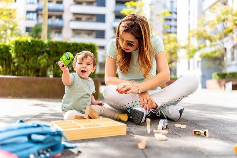 Baby boy raising a toy smiling and playing with his mother sitting on an urban park