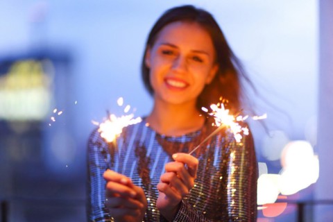 Delighted young lady in evening dress smiling and waving burning sparklers during holiday
