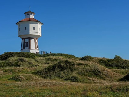A white and red lighthouse stands on a grassy dune under a cloudless blue sky, white wasertower in