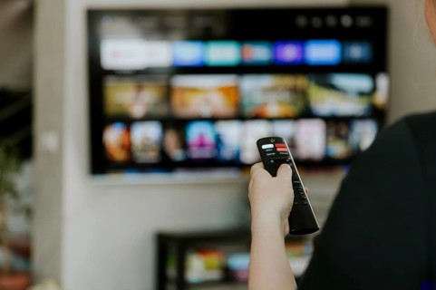 Hand holding remote control pointing to TV. Person watching smart television indoors. Blurred