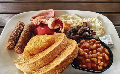 Full english breakfast on a plate, featuring fried eggs, bacon, sausages, baked beans, mushrooms,