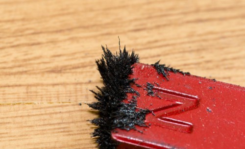 Closeup of iron filings attached to red colored north end of magnet on wood grain background