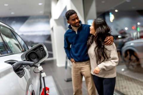 Multiethnic couple embracing while choosing a new electric car in a dealership, focusing on