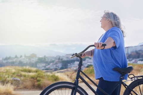 An overweight mature woman rides a black retro-style bicycle against a cloudy mountain backdrop