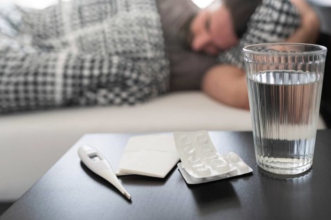 meds, medical thermometer and glass of water on nightstand against sick person suffering from flu