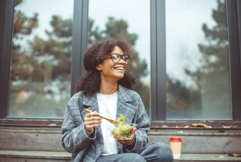 Happy african american woman office worker eating salad and smiling at camera while sitting on