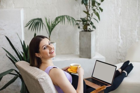 Happy pregnant young adult woman sitting on a sofa, working on a laptop and holding a cup of coffee
