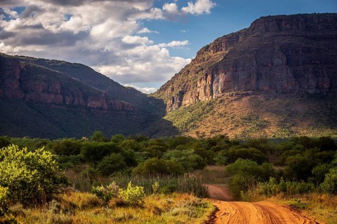 A dirt road leads through a picturesque mountain landscape with lush vegetation under a partly
