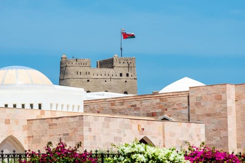 An old fortress with the Omani flag rises above pink stone buildings under a blue sky, Al Alam