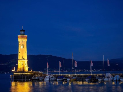 Illuminated lighthouse in the harbour at night, calm water and boats illuminated with warm light,