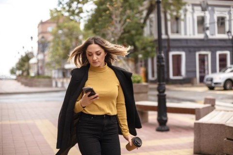 Young attractive woman with smartphone walking city street outdoors on autumn day, stylish business