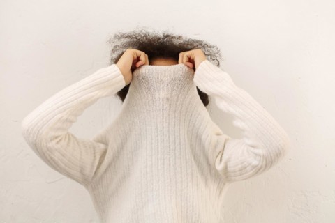 African curly female covering face with warm white sweater collar isolated on light studio