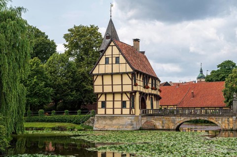 Old half-timbered house on a quiet pond with water lilies, surrounded by greenery under a cloudy