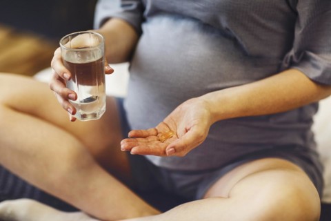Supplements during pregnancy. Young happy pregnant woman taking prenatal vitamins, holding glass of