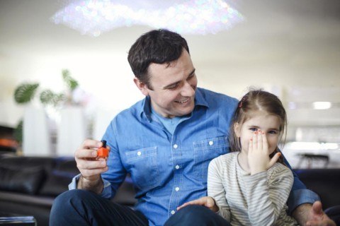 Cheerful father playing with little daughter sitting on couch and helping to paint her nails with