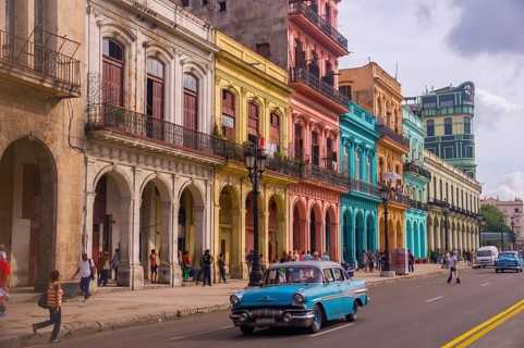 A blue oldtimer taxi is driving through Habana Vieja in front of a colorful facade of colonial