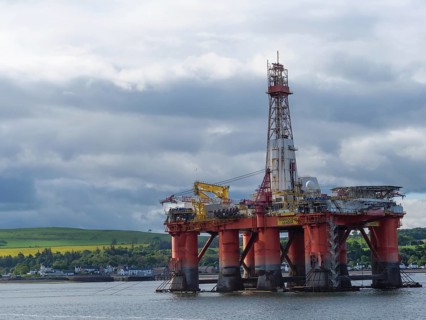 An oil drilling platform stands in the calm sea, behind it a cloudy sky and green landscape,
