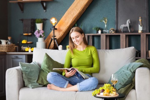 Young blond female reading interesting magazine while sitting on comfortable couch near table with