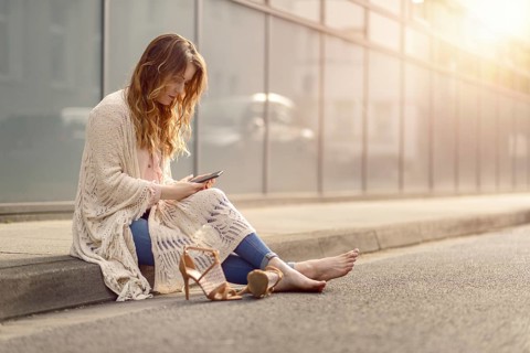 Attractive trendy woman sitting on the kerb of an urban street with her high heeled shoes lying on