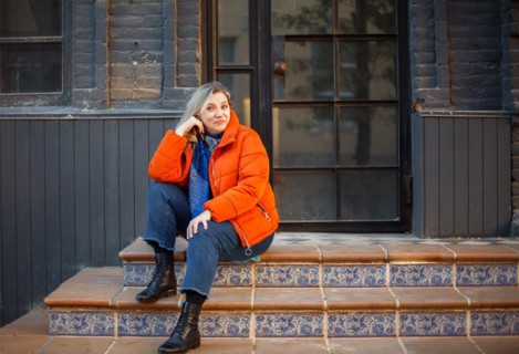 Happy smiling middle aged woman in orange down jacket sitting on concrete stairs outdoors and