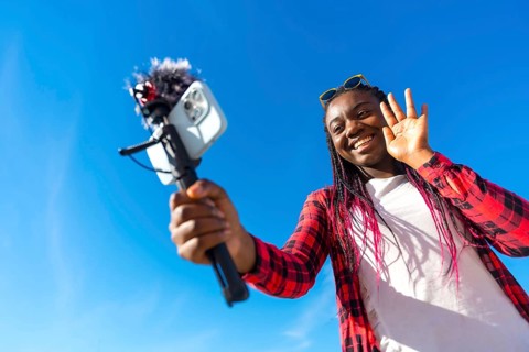 African woman waving while vlogging using mobile phone and microphone outdoors