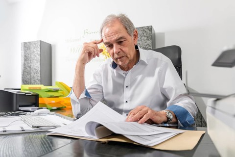 Elderly man in a white shirt sits at a desk and leafs through a document file