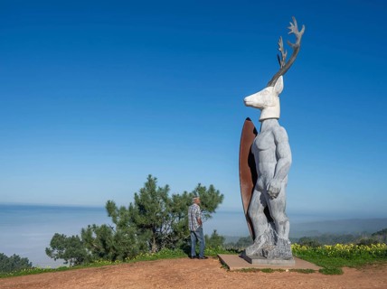 A man stands in front of a surfer statue with a deer head, Nazaré, Portugal, Europe