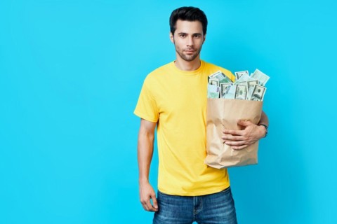 Handsome man hold paper bag full of stacks of money isolated on blue background. Success, business,
