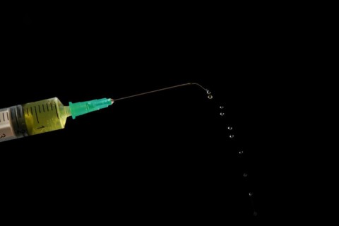 Syringe with needle, a row of drops of yellow liquid coming out, isolated on black background and
