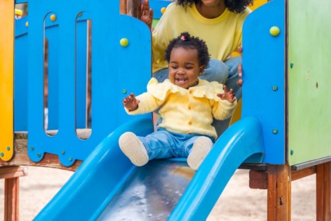 Baby girl sliding in a blue slide in a park with her mother in her back watching and controlling