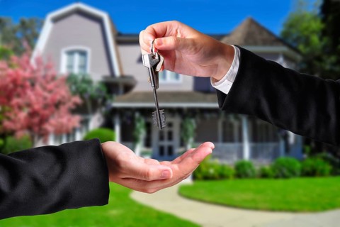 A hand giving a key to another hand. Both persons in suits and a house in the background