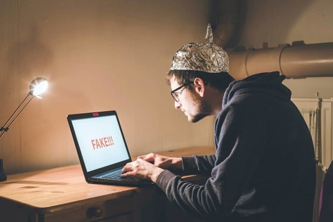 Young man with aluminum cap is sitting in the dark basement in front of a laptop. Conspiracy theory