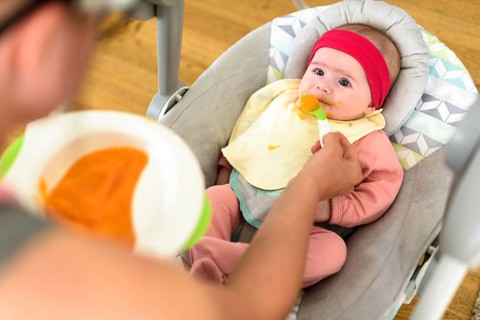 Mother feeding hungry five month old funny baby with solid food. Beagle dog watching curiously
