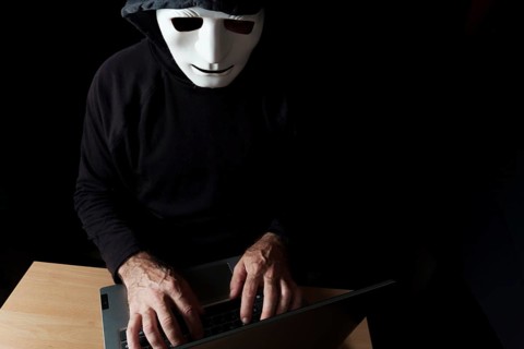 Hacker man dressed in black, hooded with mask typing on a computer cybercrime concept