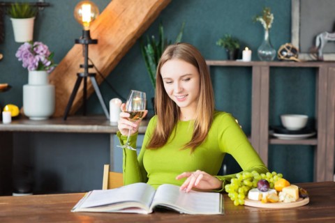 Young woman reading an interesting magazine at wooden table with fruits and wine on a weekend at