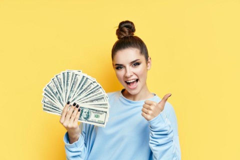 Happy excited young woman showing money banknotes with thumbs up on yellow background. Motivation,