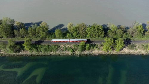 A red local railway runs on an embankment between two bodies of water with different colours, drone