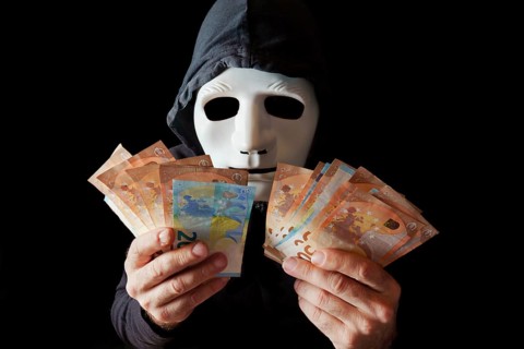 Hacker man dressed in black with white mask and black hood counting euro banknotes in his hands,