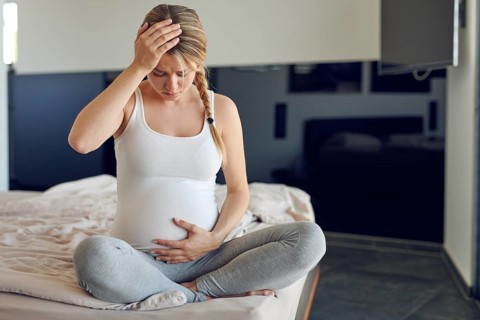 Troubled heavily pregnant young woman sitting cross legged on a bed cradling her swollen abdomen