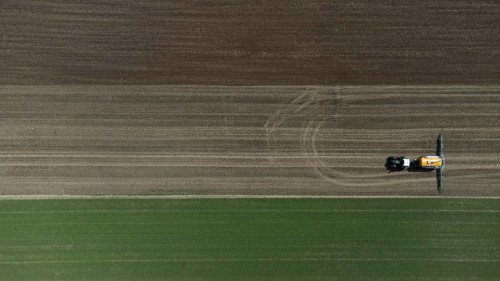 A green tractor with a yellow slurry tanker fertilises a harvested field, bird's eye view, drone