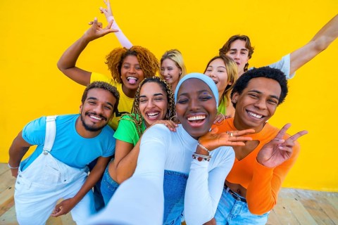 Frontal view of a multi-ethnic group of friends gesturing happily while taking a selfie in a yellow