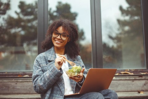 Happy african american woman eating salad, having healthy lunch while resting outdoors with laptop
