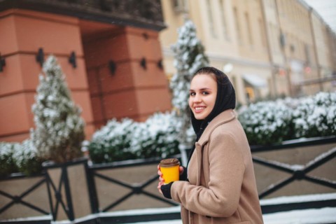 Side view of glad young female in outerwear smiling and looking at camera while enjoying takeaway