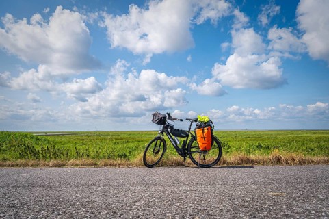 A loaded touring bike stands at the roadside with a wide field and open sky in the background,