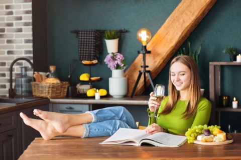 Young woman reading an interesting magazine at wooden table with fruits and wine on a weekend at
