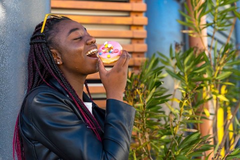 Cheerful African woman with leather jacket biting a doughnut in the corner of street
