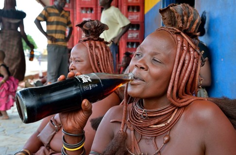 Traditionally dressed Himba woman drinking a coke in front of a bar, Opuwo, Kaokoland, Kunene,