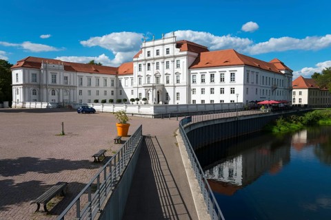 A baroque castle next to a bridge over a river, surrounded by flowering plants, Oranienburg Castle,