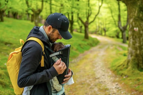 A young father with a yellow backpack walking with the newborn child in the backpack on a path in