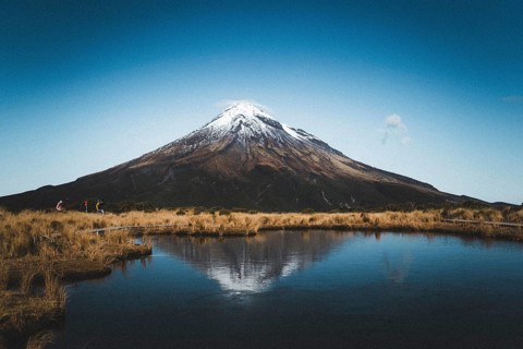 A snow-capped mountain is reflected in a still lake while people walk along the shore, Mount